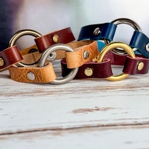 May include: Four leather bracelets with metal rings. The bracelets are in shades of brown, tan, burgundy, and blue. The metal rings are silver and gold.