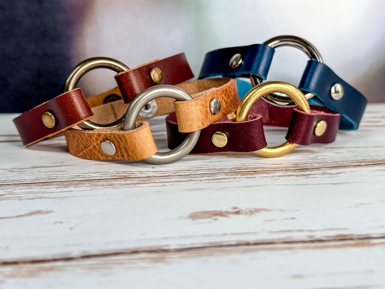 May include: Four leather bracelets with metal rings. The bracelets are in shades of brown, tan, burgundy, and blue. The metal rings are silver and gold.