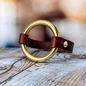 May include: A brown leather bracelet with a gold-toned metal ring. The bracelet has a snap closure.
