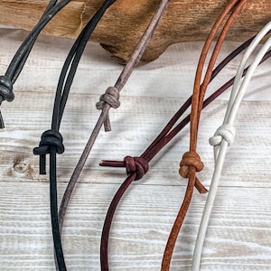 May include: Five leather cords in different colours, including black, brown, grey, and white, are tied in knots. The cords are arranged on a light wood surface.