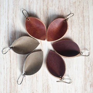 May include: Six pairs of leather earrings in various shades of brown, including tan, light brown, and dark brown. The earrings are shaped like teardrops and have a simple design.
