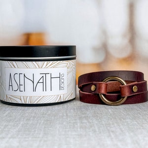 May include: A brown leather wrap bracelet with a large gold-toned ring and two silver-toned studs. The bracelet is on a white and brown textured surface. The text "ASENATH DESIGNS" is on a white tin container with a black lid.