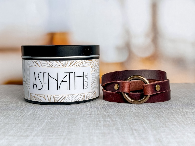 May include: A brown leather wrap bracelet with a large gold-toned ring and two silver-toned studs. The bracelet is on a white and brown textured surface. The text "ASENATH DESIGNS" is on a white tin container with a black lid.