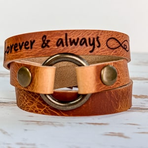 May include: A brown leather wrap bracelet with an infinity symbol and the words "forever & always" engraved on it. The bracelet has a metal ring closure.