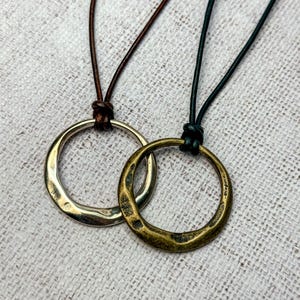 May include: Two necklaces with circular pendants. One pendant is silver-toned, the other is bronze-toned. Both pendants are attached to leather cords. The silver pendant is on a brown cord, and the bronze pendant is on a black cord. The pendants are slightly textured.