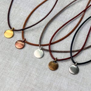 Puede incluir: Cinco collares de cordón de cuero con colgantes circulares de metal de diferentes colores. Los colgantes son de oro, cobre, plata, bronce y plata.