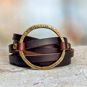Puede incluir: Un brazalete de cuero marrón con un gran acento metálico circular de color dorado. El acento metálico tiene una superficie texturizada y parece tener marcas o inscripciones.