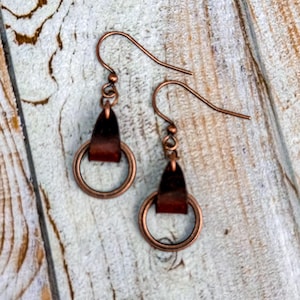 以下が含まれることがあります： 銅製のドロップイヤリングで、茶色の革のアクセントが施されています。イヤリングは、革のループがぶら下がっている小さな円形をしています。