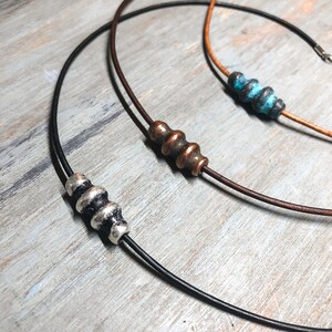 May include: Three leather necklaces with different colored beads. The necklaces are on a wooden surface. The beads are made of metal and have a textured surface.