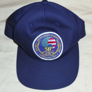 May include: Navy blue baseball cap with a commemorative patch. The patch features the words "United States of America" and "50th Anniversary of the Korean War Commemoration" with a design of the American flag and a globe.