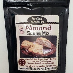 May include: A black and white bag with a label that says "Papa Rays Marketplace Almond Scone Mix". The label also shows a picture of a scone and a chef. The bag is 16 oz (454g) and makes 8-12 scones. The label says "With real almonds throughout, each bite will have you wanting more and more! Because All Mixes Are Not Created Equal."