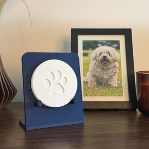 May include: A dark blue L-shaped display stand holding a circular white disc with an embossed paw print design. The stand features two small pegs at the base to support the disc, which is positioned upright for viewing on a flat surface.