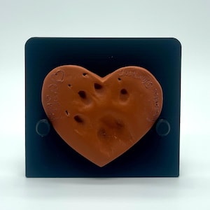 Puede incluir: Una huella de pata en forma de corazón, de color terracota, sobre un cuadrado negro. El corazón tiene una impresión de huella de pata y las palabras "In Loving Memory" y "Best" están inscritas. Dos sujetadores negros aseguran el corazón al cuadrado.
