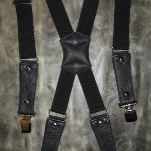 Puede incluir: Tirantes de cuero negro con clips metálicos plateados. Los tirantes tienen un patrón texturizado y están cruzados en la espalda.