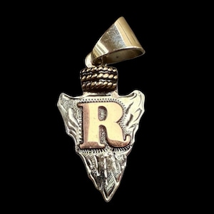 Op de afbeelding: Zilverkleurige hanger in de vorm van een pijl, met de letter "R" in goud. De hanger heeft een decoratief touwdetail en een zilveren beugel voor een ketting. De achtergrond is zwart.