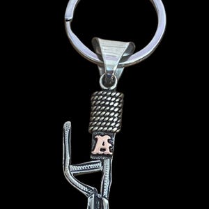 Puede incluir: Llavero plateado con un dise&ntilde;o decorativo y la letra "A" dorada. El llavero tiene un anillo partido para llaves y un elemento decorativo con textura de cuerda. La letra "A" est&aacute; en una fuente serif.
