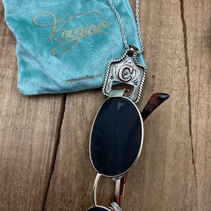 May include: A silver pendant necklace with a decorative frame and a letter "C" in the centre. The pendant rests on sunglasses with dark lenses. A teal velvet bag with gold lettering is in the background.