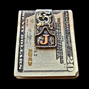 May include: A decorative money clip with a gold and silver filigree design, featuring the letter "J". The clip is holding a stack of twenty-dollar bills. The clip has a rope-like border and a gold-colored initial.