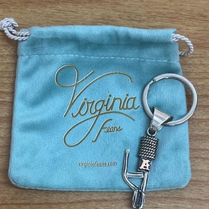 Puede incluir: Un llavero plateado con un dise&ntilde;o decorativo y la letra "A" en un tono oro rosado. El llavero est&aacute; junto a una bolsa de terciopelo azul claro con las palabras "Virginia Fears" y la direcci&oacute;n del sitio web impresas en dorado.