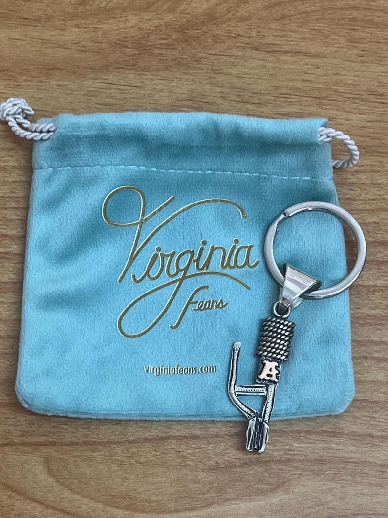 Puede incluir: Un llavero plateado con un dise&ntilde;o decorativo y la letra "A" en un tono oro rosado. El llavero est&aacute; junto a una bolsa de terciopelo azul claro con las palabras "Virginia Fears" y la direcci&oacute;n del sitio web impresas en dorado.