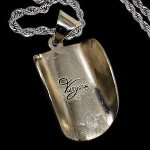 May include: A silver-colored pendant with the word "Virginia" in cursive script. The pendant is attached to a silver chain. The pendant has a curved, shovel-like shape.