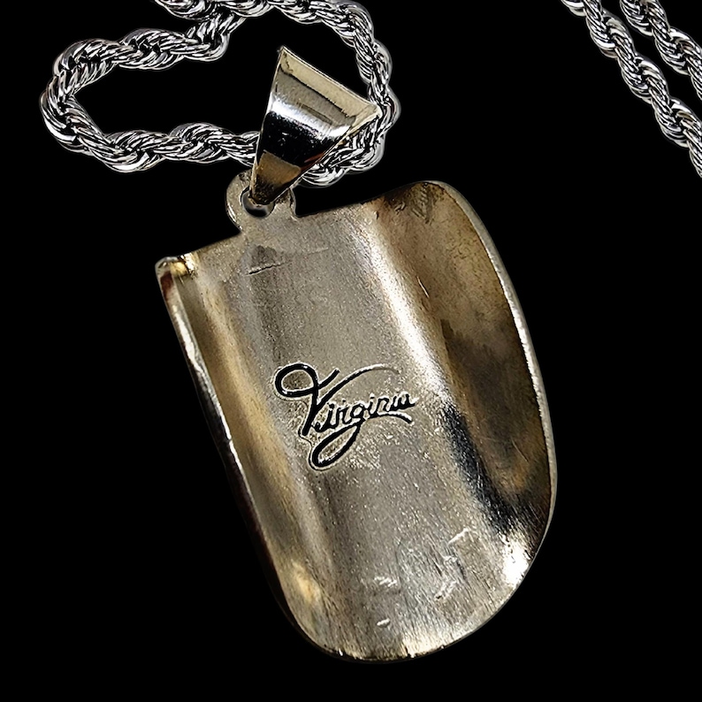May include: A silver-colored pendant with the word "Virginia" in cursive script. The pendant is attached to a silver chain. The pendant has a curved, shovel-like shape.