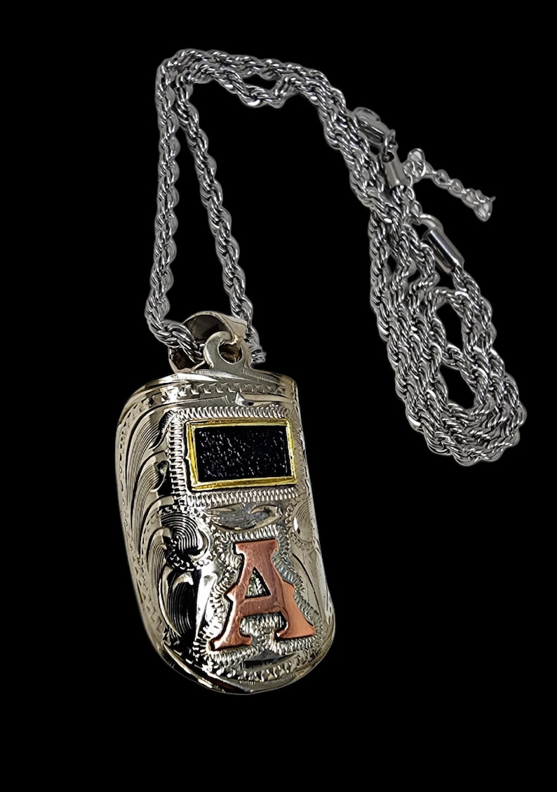 May include: Silver-tone pendant necklace featuring a detailed, engraved design. The pendant has a rectangular black and gold accent and a copper-colored letter "A". The necklace has a rope chain.