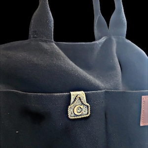 Puede incluir: Bolso tote de lona negra con un bolsillo frontal y un cierre decorativo de metal dorado. El cierre presenta un diseño de cuerda y la letra "C" dorada. El bolso tiene dos asas negras.