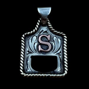 May include: Silver pendant featuring the letter "S" in copper. The pendant has a decorative border and a bail for a chain. The design includes floral elements and a rope-like trim, suitable for a necklace. The pendant is approximately 3 cm in height.