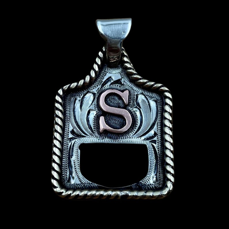 May include: Silver pendant featuring the letter "S" in copper. The pendant has a decorative border and a bail for a chain. The design includes floral elements and a rope-like trim, suitable for a necklace. The pendant is approximately 3 cm in height.