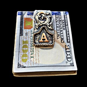 May include: A money clip with an ornate gold and silver design, featuring the letter "A" in the center. The clip is holding a stack of U.S. one hundred dollar bills. The background is solid black.