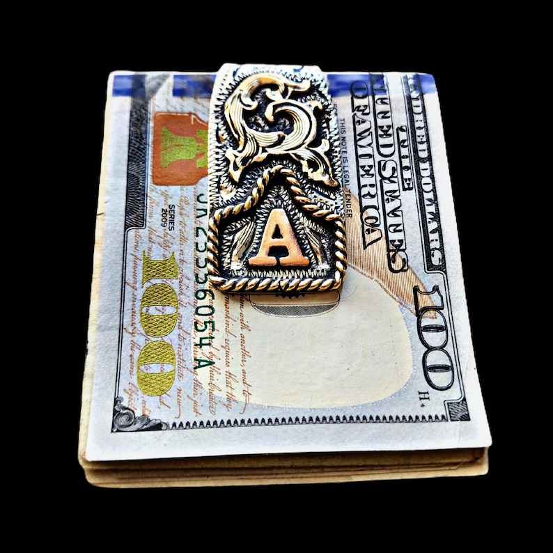 May include: A money clip with an ornate gold and silver design, featuring the letter "A" in the center. The clip is holding a stack of U.S. one hundred dollar bills. The background is solid black.