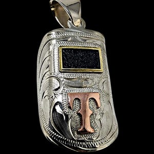 May include: Silver pendant in the shape of a welding mask, featuring intricate engraved patterns. The pendant has a gold-colored frame around a black rectangle and a copper-colored letter "T". The bail is silver.