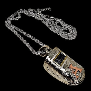 May include: Silver-toned pendant necklace with a detailed, engraved design. The pendant features a rectangular black stone with a gold border and a copper-colored letter "A". The chain is a silver rope style.