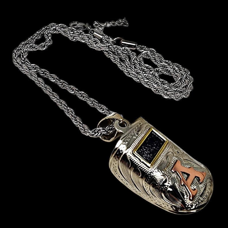 May include: Silver-toned pendant necklace with a detailed, engraved design. The pendant features a rectangular black stone with a gold border and a copper-colored letter "A". The chain is a silver rope style.