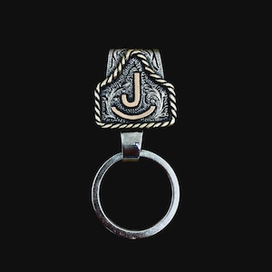 May include: Silver-toned key ring with a decorative top featuring the letter "J". The top has a floral design with gold-colored rope-like trim. The key ring is suspended from a small, rectangular connector.