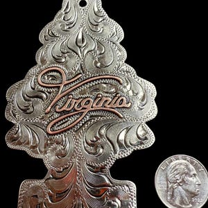 Peut inclure: Ornement en m&eacute;tal argent&eacute; en forme de sapin de No&euml;l, avec des volutes florales complexes et le mot "Virginia" en script cuivr&eacute;. Une pi&egrave;ce de monnaie am&eacute;ricaine est &agrave; c&ocirc;t&eacute; de l'ornement pour la comparaison de taille.