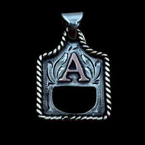 May include: Silver pendant with a decorative rope-like border. The pendant features a stylized letter "A" in a copper colour, set against an engraved floral pattern. The background is black.
