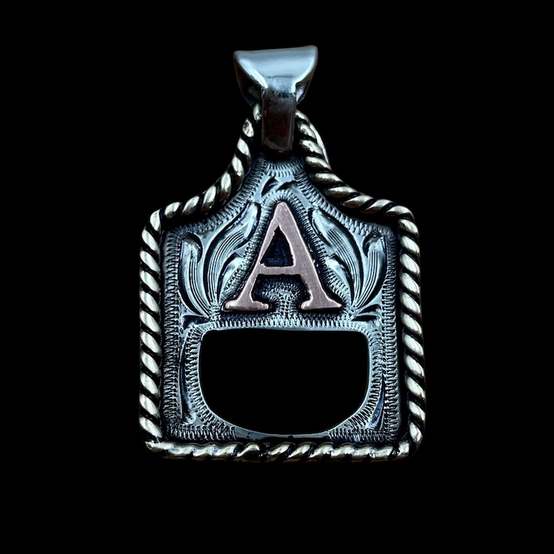 May include: Silver pendant with a decorative rope-like border. The pendant features a stylized letter "A" in a copper colour, set against an engraved floral pattern. The background is black.