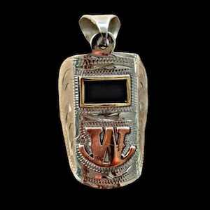 May include: Silver-toned pendant in the shape of a welding mask. The pendant features a rectangular black center framed in gold, and the letter "W" in copper-colored metal. Intricate engraved details and a loop for a chain complete the design.