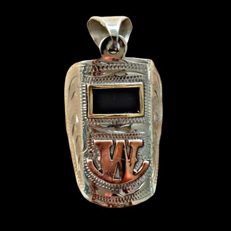 May include: Silver-toned pendant in the shape of a welding mask. The pendant features a rectangular black center framed in gold, and the letter "W" in copper-colored metal. Intricate engraved details and a loop for a chain complete the design.