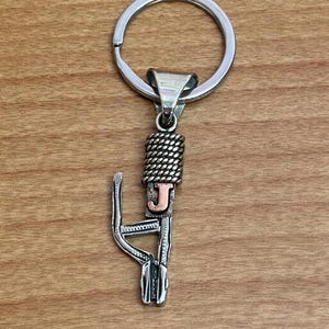 Puede incluir: Llavero de metal plateado con un dise&ntilde;o decorativo. El llavero presenta un anillo circular, una secci&oacute;n trenzada y una letra "J" estilizada en color cobre. El dise&ntilde;o est&aacute; unido a un elemento decorativo calado.
