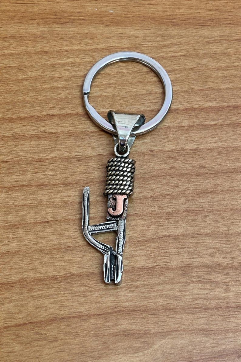 Puede incluir: Llavero de metal plateado con un dise&ntilde;o decorativo. El llavero presenta un anillo circular, una secci&oacute;n trenzada y una letra "J" estilizada en color cobre. El dise&ntilde;o est&aacute; unido a un elemento decorativo calado.
