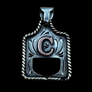 May include: Silver pendant with a decorative border and a copper-coloured letter "C" in the centre. The pendant has a rectangular shape with a rope-like border and a bail for a chain. The background is black.