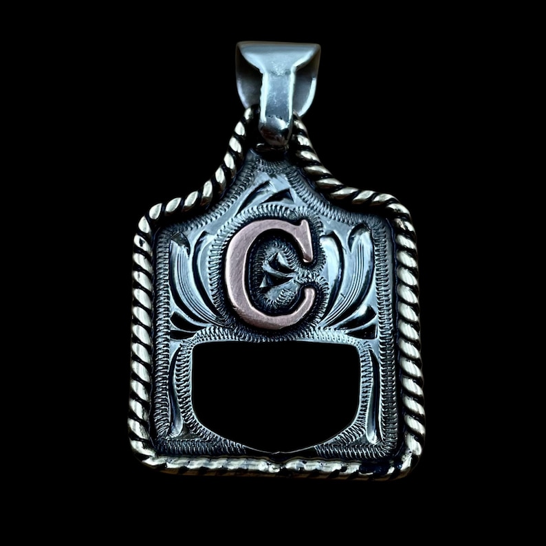 May include: Silver pendant with a decorative border and a copper-coloured letter "C" in the centre. The pendant has a rectangular shape with a rope-like border and a bail for a chain. The background is black.