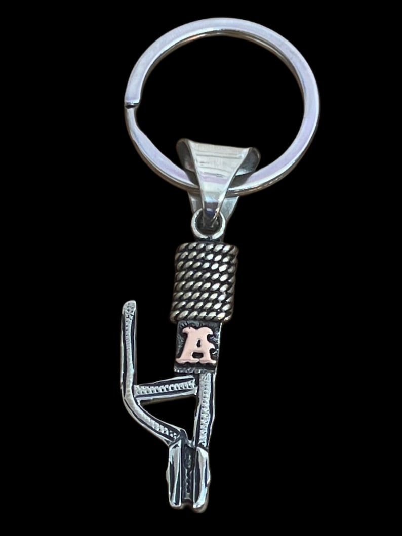 Puede incluir: Llavero plateado con un dise&ntilde;o decorativo y la letra "A" dorada. El llavero tiene un anillo partido para llaves y un elemento decorativo con textura de cuerda. La letra "A" est&aacute; en una fuente serif.