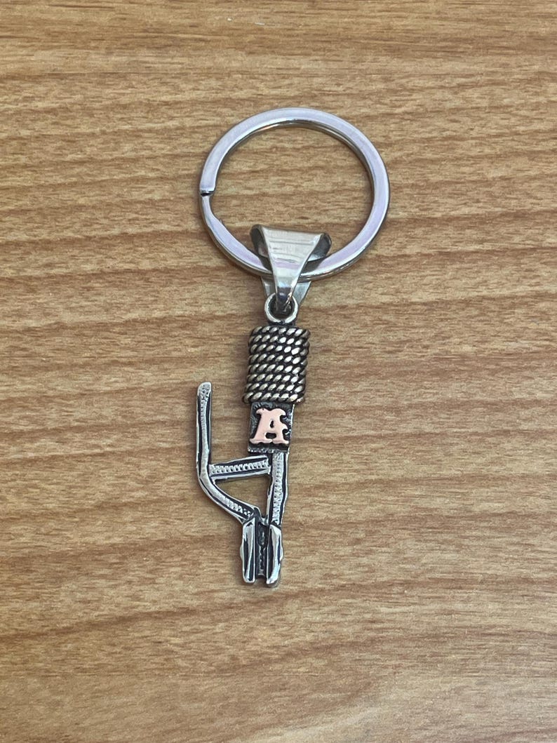 Puede incluir: Llavero de metal plateado con un anillo, una secci&oacute;n trenzada y un elemento decorativo con la letra "A". El llavero est&aacute; dise&ntilde;ado con una apariencia detallada y texturizada.