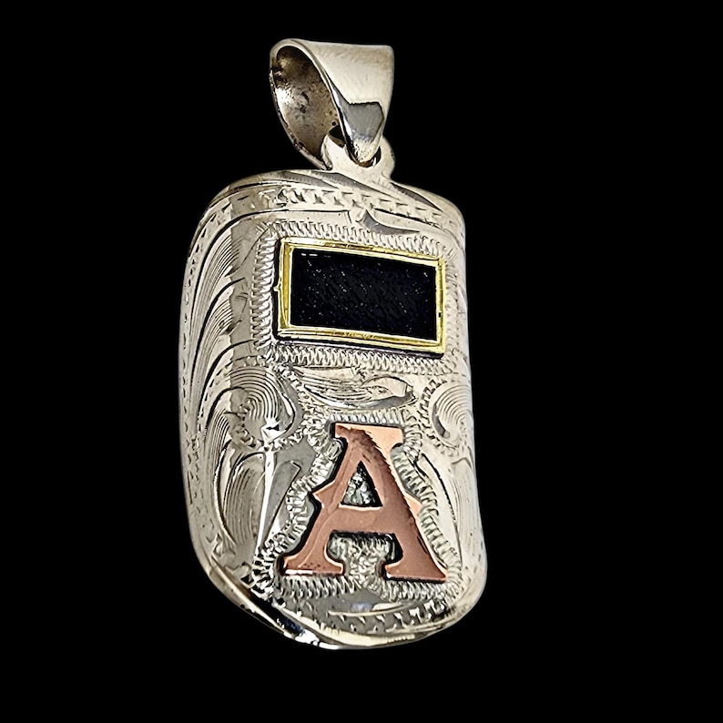 May include: Silver pendant in the shape of a welding mask, featuring an engraved floral design. The pendant has a rectangular black insert framed in gold and a copper-colored letter "A" at the bottom. The bail is silver.
