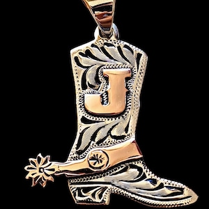 Puede incluir: Colgante dorado con forma de bota de vaquero con espuela. La bota presenta un diseño floral grabado y la letra "J" dorada. El colgante cuelga de una argolla dorada.