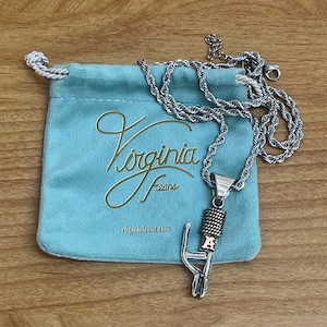 Puede incluir: Un collar con colgante de micr&oacute;fono plateado con una cadena de cuerda descansa sobre una bolsa de terciopelo azul claro. La bolsa tiene letras doradas que dicen "Virginia Feans" y la direcci&oacute;n del sitio web. El collar est&aacute; sobre una superficie de madera.
